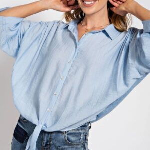 Woman in a loose light blue button-up shirt and jeans, smiling with hands near her face.