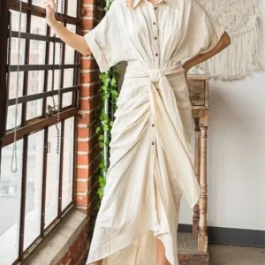 Elegant woman in a ruched white dress standing by a window.