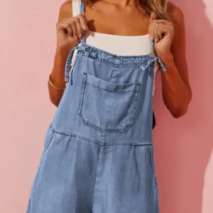 Woman wearing light blue denim overalls with a white top against a pink background.