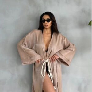 Woman posing confidently in a beige robe with sunglasses against a gray wall.